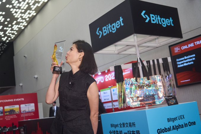 The image features a woman standing next to a display at an event. She is holding a trophy and appears to be celebrating her achievement. The background of the photo shows several other people present at the same event, indicating that this is a gathering or exhibition for various businesses and industries.