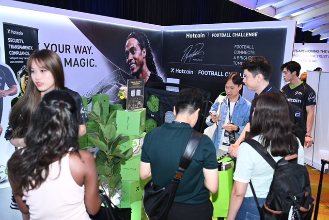 A busy exhibition booth for 'Hotcoin' advertises 'SECURITY. TRANSPARENCY. COMPLIANCE.' and a 'FOOTBALL CHALLENGE', with staff interacting with attendees.