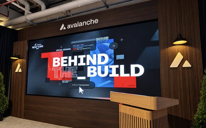 A wooden podium stands before a large display screen at the Hudson Stage during Avalanche's "Behind the Build" demo series at the New York Summit, featuring an "Initiate Transaction" button and code snippets. The Avalanche logo is prominently displayed above the screen, flanked by sconce lighting in a modern, industrial-styled venue.