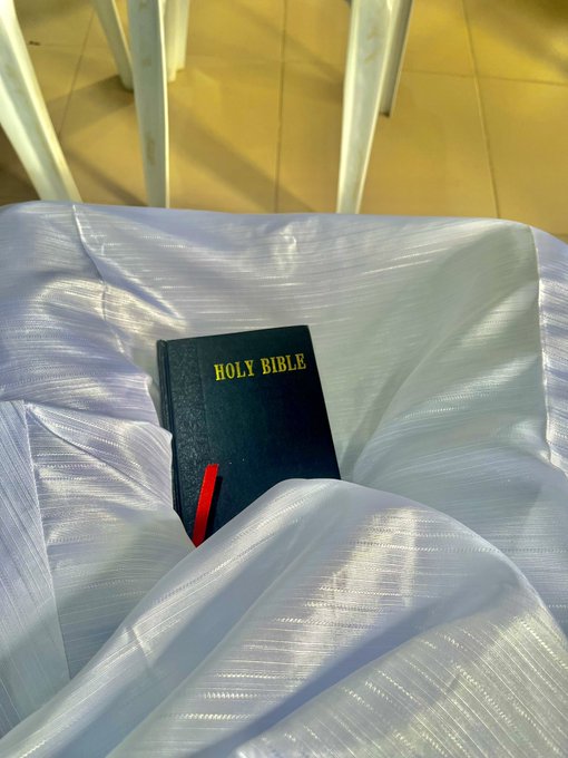 A black 'HOLY BIBLE' with a red ribbon bookmark rests on a white fabric surface, likely draped over a church pew.