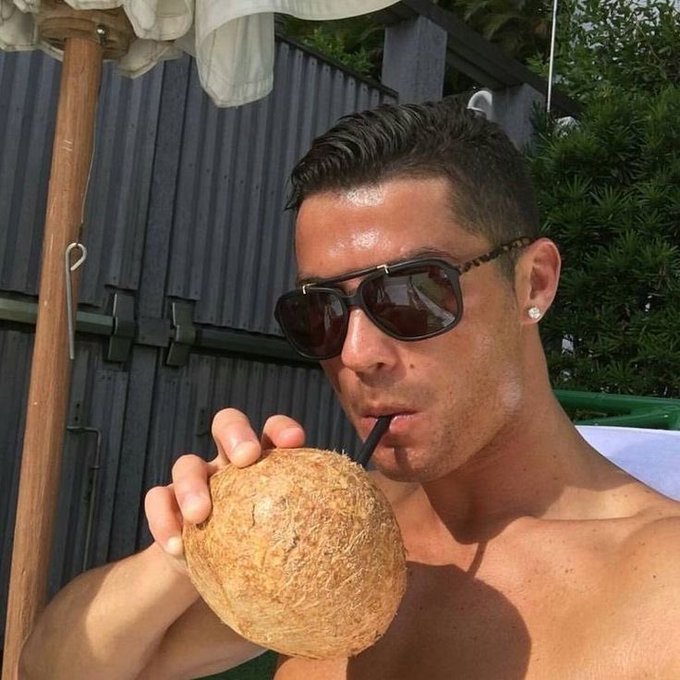 A man in sunglasses sipping from a coconut with a straw in an outdoor setting.