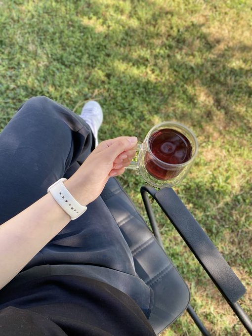 A first-person view of someone sitting outdoors on a chair, holding a glass mug of dark red tea, wearing a white smartwatch and dark pants, with grass visible below.