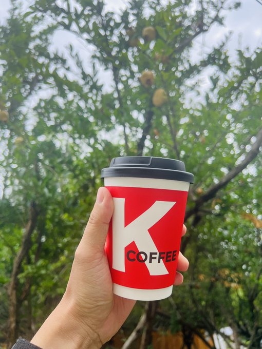 A hand holds a red and white 'K COFFEE' paper cup with a black lid, set against a backdrop of green foliage.