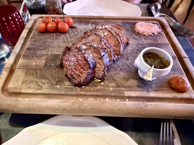 The image shows a large, thick cut steak sitting on a wooden cutting board accompanied by various condiments and sauce containers. The cutting board is placed on top of a dining table with plates, silverware, and additional food items arranged around it. In the background, there are chairs and a table, suggesting that this meal is taking place in an elegant setting or restaurant.
The image contains several details about the steak, such as its thickness (thicker than a quarter), the accompanying condiments and sauce containers, and the overall presentation of the dish.