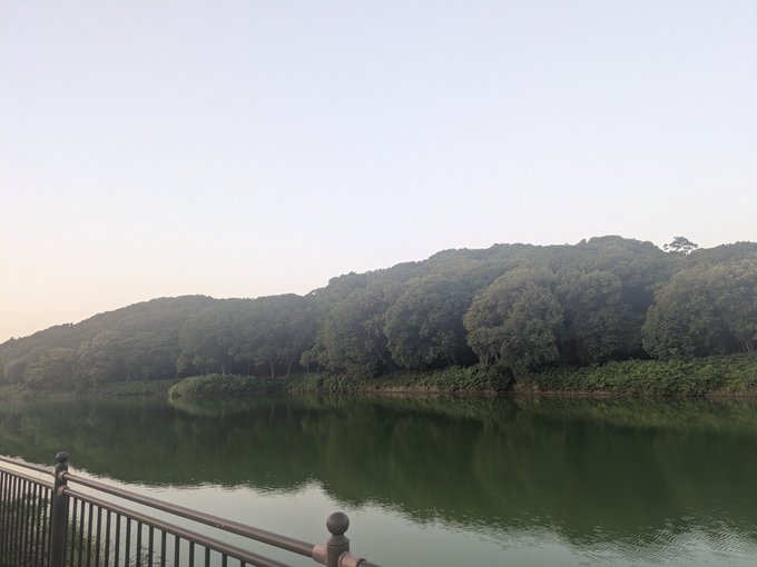 A tranquil scene of a calm green lake surrounded by forested hills, with a metal railing visible in the foreground.