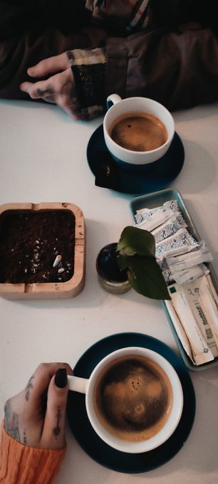 A moody overhead shot of two people sharing coffee at a white table, with their tattooed hands visible holding cups, alongside a small potted plant, a wooden planter with soil, and a tin of sugar packets.