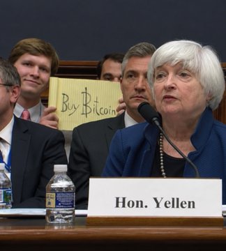 Photo of Janet Yellen at a congressional hearing, with a man behind her holding up a yellow 'Buy Bitcoin' sign.