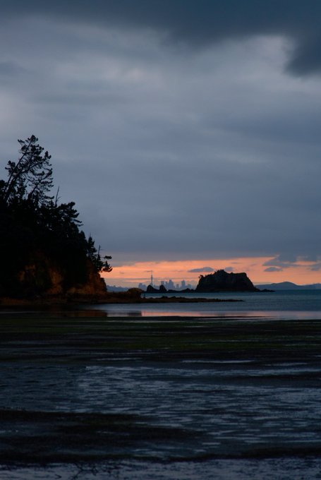 A serene coastal scene at dusk or dawn, with a dark, cloudy sky above and an orange glow on the horizon, featuring silhouetted trees on the left and rock formations in the water.