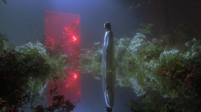 An artistic, moody image showing a woman in a white dress standing in a lush garden filled with white flowers, facing a glowing red rectangular portal reflected in still water beneath her.