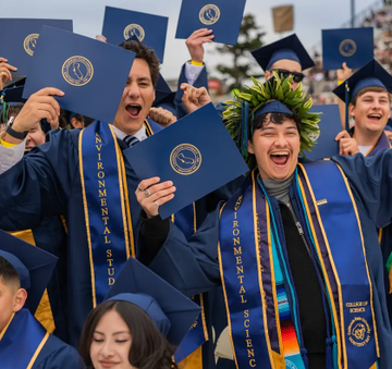 A joyous group of graduates in blue caps and gowns, wearing stoles that read "ENVIRONMENTAL STUDIES," cheer and hold up diplomas, celebrating their college achievement. The image reflects the post's question about choosing between an Ivy League education and alternative financial paths, symbolizing the traditional college graduation milestone being weighed against holding cryptocurrency.