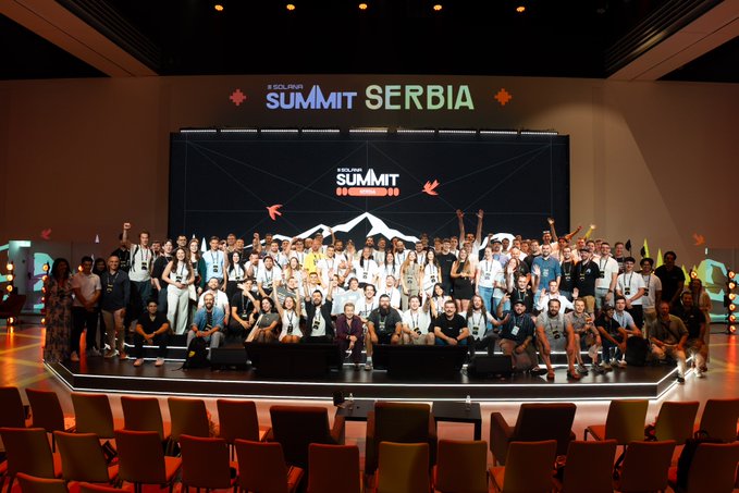 A large group photo of attendees at the "SOLANA SUMMIT SERBIA" conference, with participants posing on stage beneath the event branding displayed on a large screen.