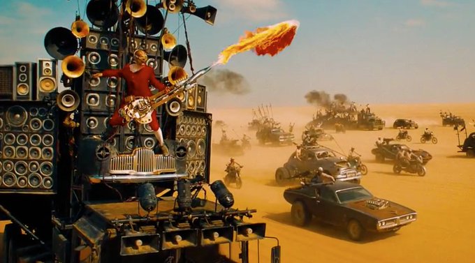 A lone guitarist in red stands atop a massive wall of amplifiers, shooting a stream of fire into the desert sky as a vast armada of post-apocalyptic vehicles speeds across the sand dunes behind him. The image humorously captures the chaos of the 2021 NFT gas wars, where the "Doof Warrior" on a wall of speakers is overwhelmed by the oncoming hoard — mirroring the intense competition and absurd energy of trying to mint NFTs while transaction fees skyrocketed.