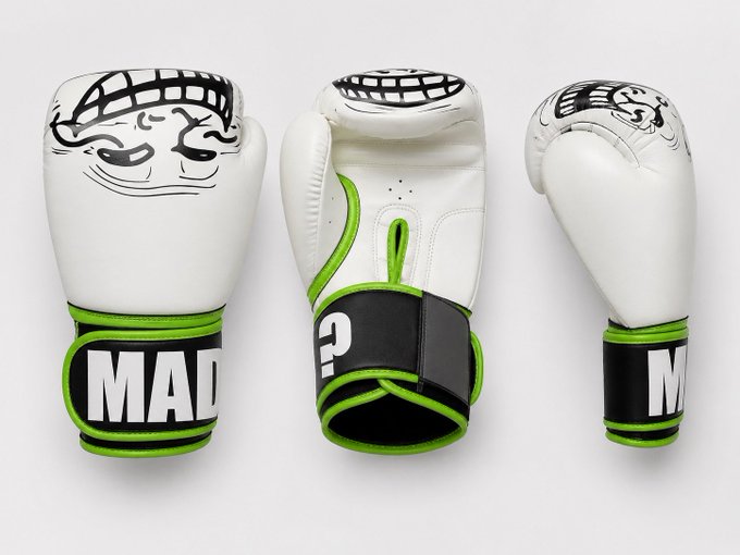 Three views of a white boxing glove with green trim, black wrist straps displaying the letters "MAD" or "M", and a stylized face logo printed on the knuckle area.