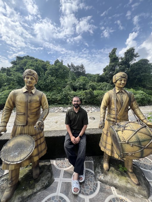 A person sitting on a wall outdoors between two golden statues of female drummers, with a forested area and river in the background.