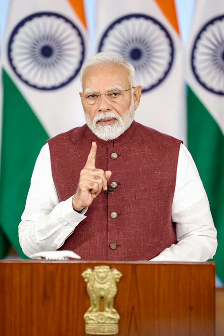Indian Prime Minister Narendra Modi speaks at a podium with the Ashoka emblem, backed by Indian flags.