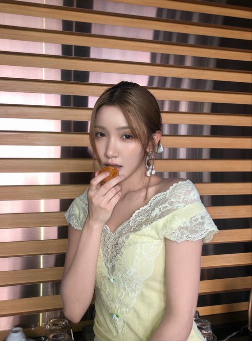 A woman in a pale yellow lace top and pearl earrings poses thoughtfully against a wooden slatted backdrop while taking a delicate bite of what appears to be a mooncake, fitting the caption's "mooncake, but make it LV" theme that elevates the traditional treat into a luxurious moment.
