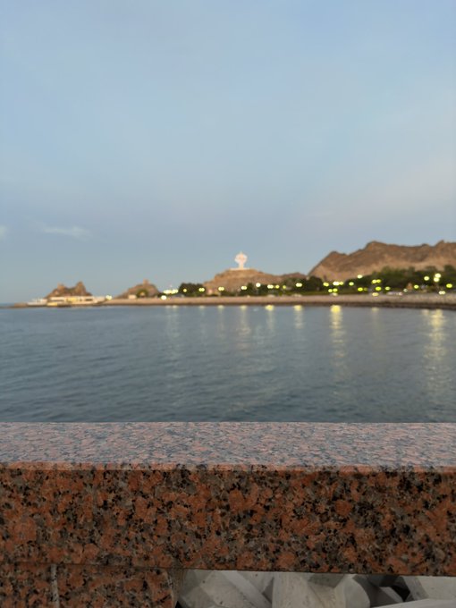 A scenic view across calm water at dusk toward a lit coastal promenade with mountains in the background, framed by a granite ledge in the foreground.