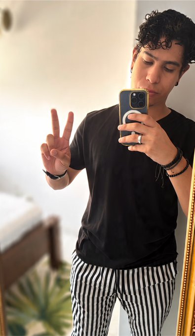 A young man with curly dark hair taking a mirror selfie in a bedroom, wearing a black t-shirt, black-and-white striped pants, and a phone case with a ring holder, making a peace sign.