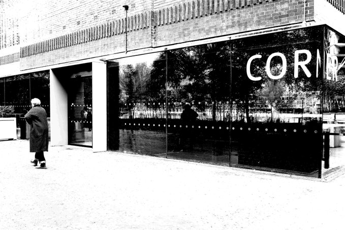 A black and white street photograph showing a person in a long coat walking past a building with large reflective black windows displaying the partial text 'CORN'.