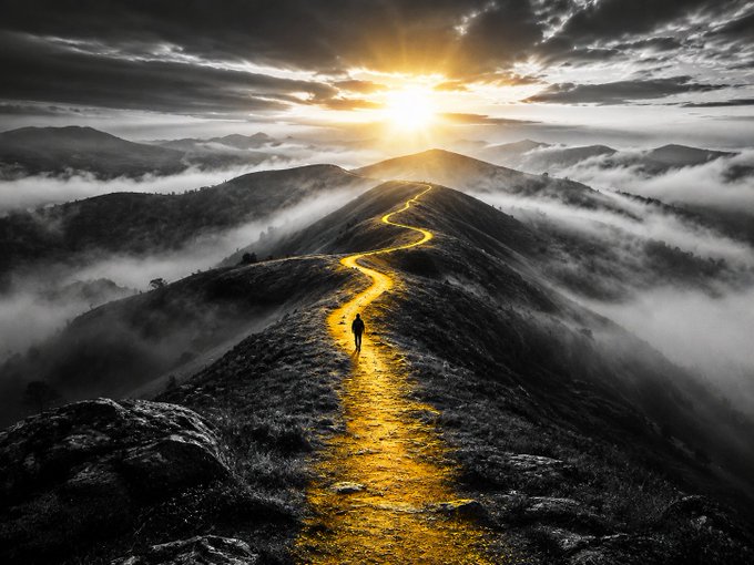 A dramatic black-and-white landscape with a winding golden path along a mountain ridge leading toward a bright sun, where a lone figure walks amid clouds and mist.