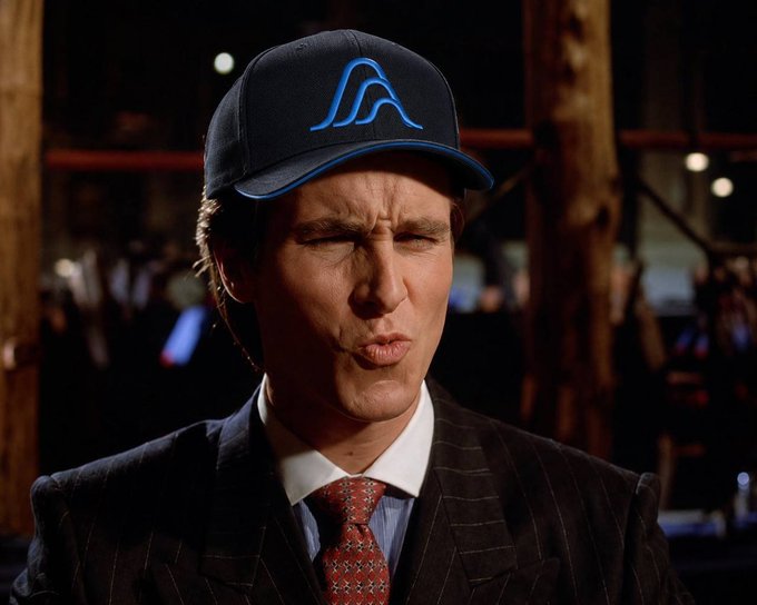 The caption relates to the image:

A man in a pinstripe suit and red patterned tie puckers his lips, wearing a dark baseball cap with a blue "Λ" logo, as he waits for the "Variational" debut in October.

Caption: A man puckers his lips while wearing a dark cap featuring a blue "Λ" logo, dressed in a pinstripe suit and red patterned tie.