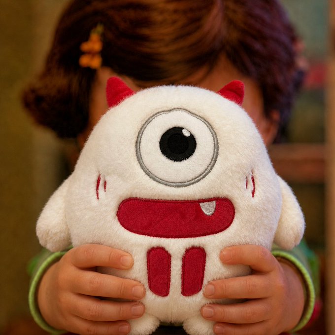 A young child holds up a fluffy white monster plush toy with a single large eye, red horns, and a red mouth with one tooth, partially obscuring their face.