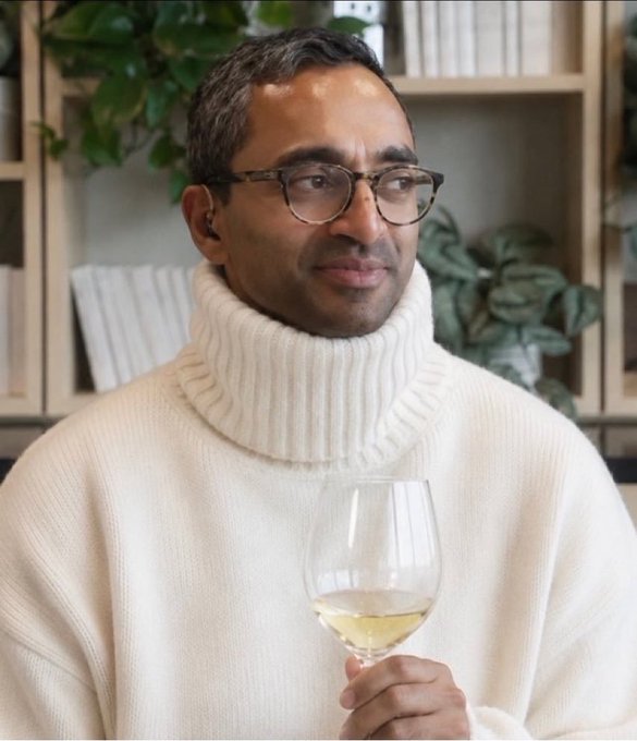 A man wearing glasses and a cream-colored turtleneck sweater holds a glass of white wine and smiles slightly indoors.