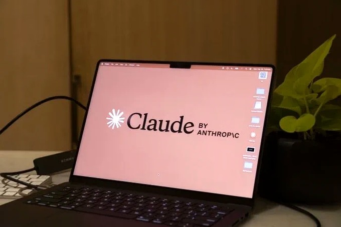 A photograph of a modern laptop open on a wooden desk, displaying a pink screen with the 'Claude by ANTHROPIC' logo in black text. A small green potted plant and part of a keyboard are visible next to the laptop against a wood-paneled background.
