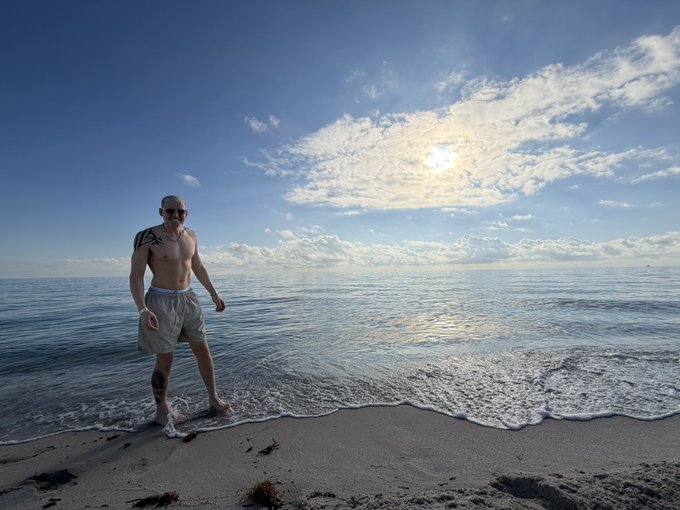 A shirtless man with tattoos and sunglasses stands barefoot at the ocean's edge on a sandy beach under a partly cloudy sky, smiling at the camera.

The relaxed beachside pose complements the post's motivational message about seizing opportunities ("AntHood GTD seats are open") by suggesting a carefree, "life's a beach" mindset of someone ready to take a winning chance.