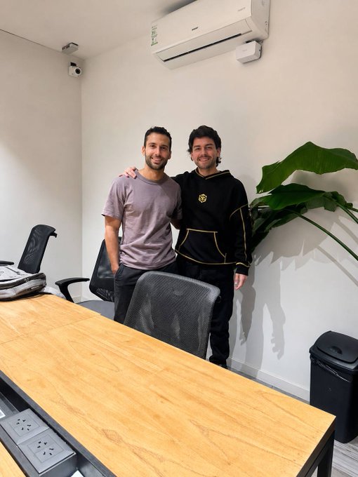 The image features two men standing next to each other in an office setting. They are both smiling for a photo, showcasing their camaraderie or perhaps celebrating a recent accomplishment. The room has several chairs placed around the area, indicating that this is likely an office environment. A potted plant can be seen on one side of the room, adding some greenery to the space. In addition to these elements, there are also two backpacks and a handbag visible in the scene, suggesting that the men might have been working together or spending time at the office recently.