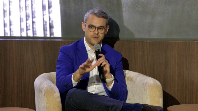 A man with glasses and a blue blazer speaking into a microphone while seated in a beige armchair during what appears to be a panel discussion or interview.