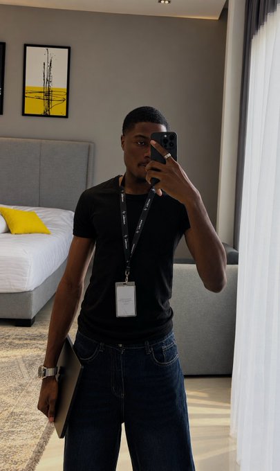 A man in a black t-shirt and jeans takes a mirror selfie with his phone in a hotel-style room, holding a MacBook in one hand and wearing a lanyard with an ID badge, likely on a business trip or work assignment.