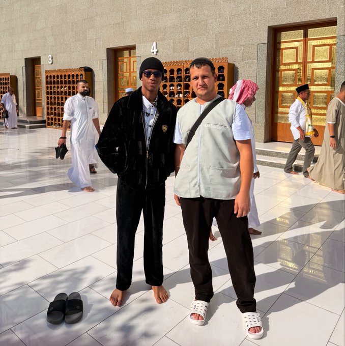 Two men stand barefoot in front of ornate golden doors marked "3" and "4" at what appears to be a religious site, with the taller man on the left wearing a black beanie and tracksuit while the other wears a vest labeled "LEGEND" and holds sandals. The caption "gm if u gm back" playfully nods to the photo's iconic pairing of casual street style against a sacred backdrop, suggesting an easy, good-morning-style rapport between the two friends.