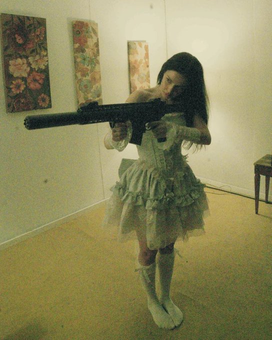A young woman with long dark hair wearing a frilly, light-colored dress stands in a room aiming a large black firearm toward the camera.