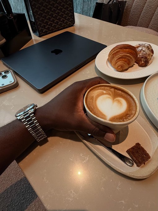 A first-person view of someone holding a cappuccino with beautiful latte design, with a plate of baked goods including croissants and a brownie on the marble table, alongside a closed MacBook laptop and iPhone.