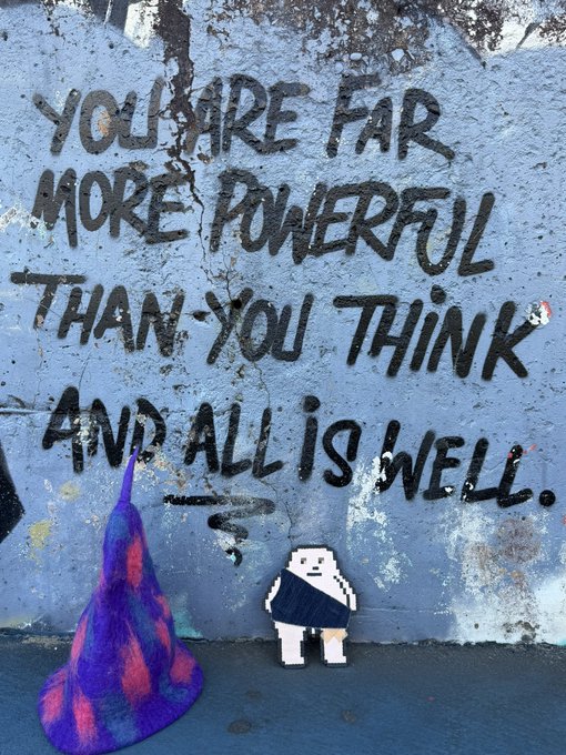 Street art mural reading 'YOU ARE FAR MORE POWERFUL THAN YOU THINK AND ALL IS WELL.' painted on a blue wall, with a small paper figure and purple sculpture at the base.