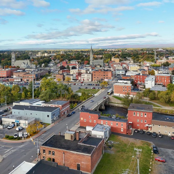 An aerial view of downtown Plattsburgh, New York, showcasing its historic brick buildings, church spires, and a bridge crossing a river, reflecting the small-city backdrop where local officials are now considering a 12-month moratorium on new large-scale crypto mining and AI data center projects consuming 300kW or more.