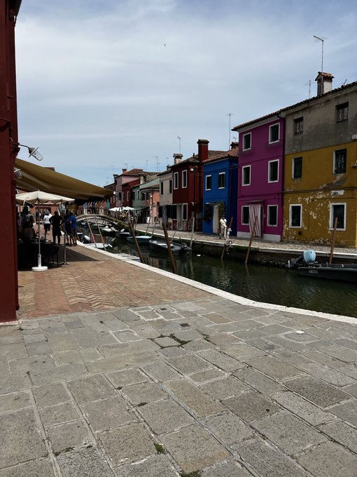 A view of a canal in Burano, Italy, with colorful houses (yellow, blue, magenta) lining the water and a stone walkway in the foreground.