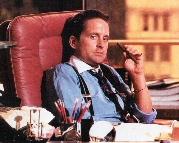 The image features Michael Douglas as the iconic character Gordon Gekko from the 1987 film "Wall Street," shown in his signature power pose—leaning in a red leather office chair, wearing suspenders and a loosened tie, holding a cigar with a confident smirk. The caption playfully casts Gekko as a crypto market mover, humorously suggesting that only this legendary "greed is good" financier can take a token from zero to a $200M market cap.