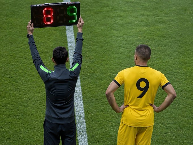 A soccer official holds up the substitution board displaying "8 9" on the sideline next to player #9 in a yellow jersey wearing the number 9 on his back, aligning with the post's tribute to Binance's 9th anniversary by symbolizing a "number 9" theme.