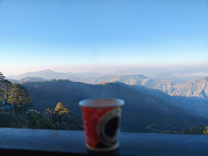 A red paper cup sits on a ledge overlooking a scenic mountain range with rolling forested hills under a clear blue sky at dawn.