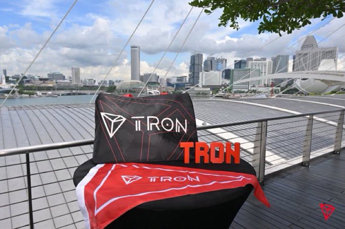 A black TRON-branded item with red accents sits on a railing overlooking a city waterfront with modern skyscrapers and a bridge in the background.