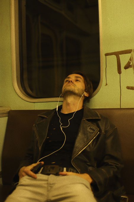 A man in a leather jacket relaxes on a subway or train seat with his eyes closed, listening to music through white wired earbuds.