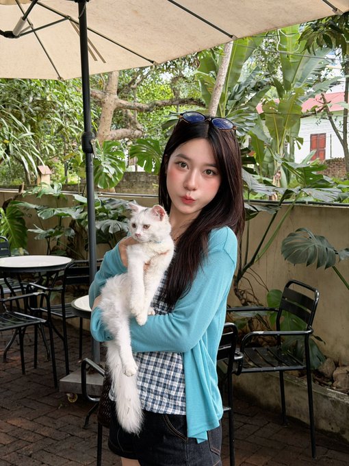 A young woman with long dark hair holds a white cat outdoors under a patio umbrella, surrounded by lush green tropical plants.