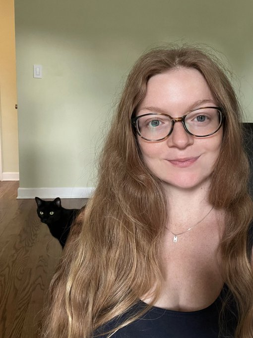A woman with long auburn hair and glasses smiles softly at the camera while her black cat, Nox, photobombs the background with bright yellow eyes peeking around the corner. The sage green room and warm wooden floor create a cozy home setting for this feline guest appearance.