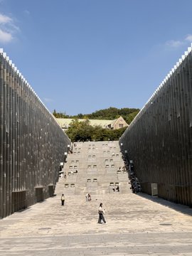 A grand outdoor staircase flanked by tall slatted walls leads up to a traditional Japanese building with a green tiled roof, with small figures of people walking on the steps under a clear blue sky.