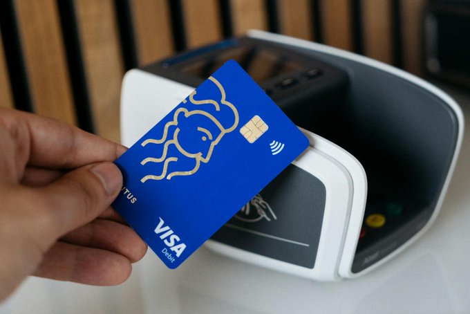 A hand holds a blue Visa debit card with a gold mermaid design near a white payment terminal.