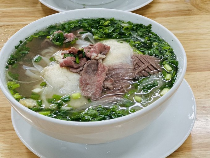 A bowl of Vietnamese pho noodle soup containing beef slices and herbs, served on a white plate on a wooden table.