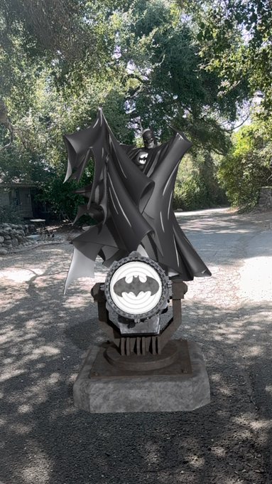 A Batman statue with a flowing cape and the Bat-symbol on its chest is displayed outdoors on a concrete base.