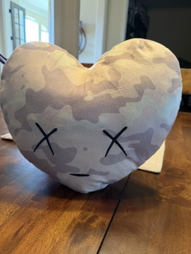 A heart-shaped plush toy with a gray camouflage pattern and X-shaped eyes sitting on a wooden floor.